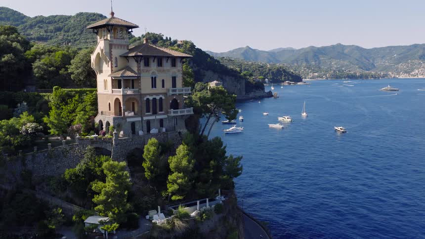 Aerial view flying towards a luxurious historic villa perched on a cliffside overlooking the beautiful blue waters of Portofino, Italy