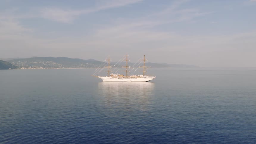 An aerial shot slowly approaches a magnificent white tall ship anchored in the calm blue sea with a distant, hazy coastline