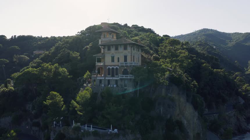 Drone shot pulls back to reveal a stunning Italian villa built on a cliffside amidst a lush green forest at sunset