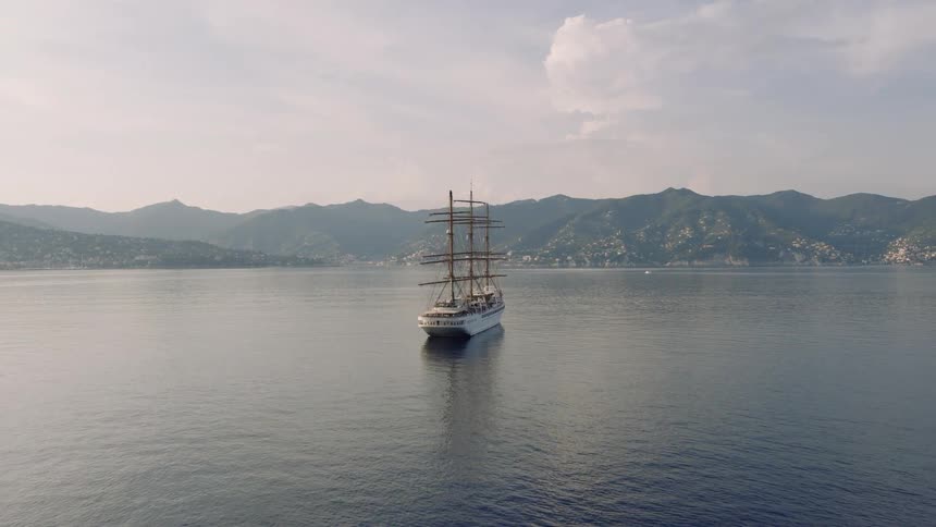 A majestic three-masted tall ship is anchored in a tranquil bay as the camera slowly pulls back to reveal the mountainous coastline