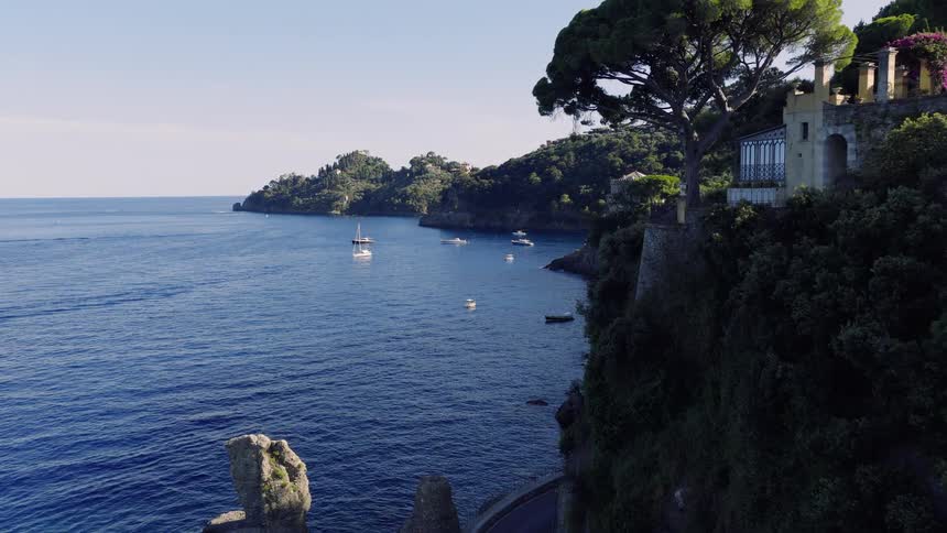 A breathtaking aerial view captures the scenic road winding along the lush, rocky coastline of the Italian Riviera, with boats dotting the tranquil blue sea