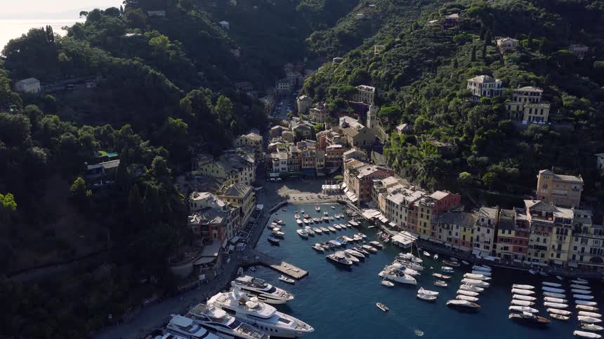 A stunning aerial flight reveals the picturesque harbor of Portofino, Italy, filled with luxury yachts and surrounded by colorful buildings and lush green hills