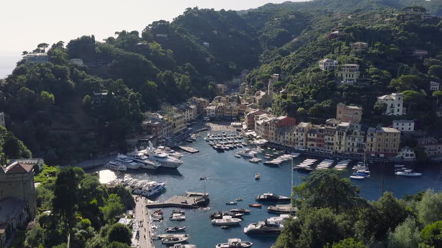 Stunning aerial view flying over lush green hills to reveal the iconic harbor of Portofino, Italy, with its colorful houses and docked yachts
