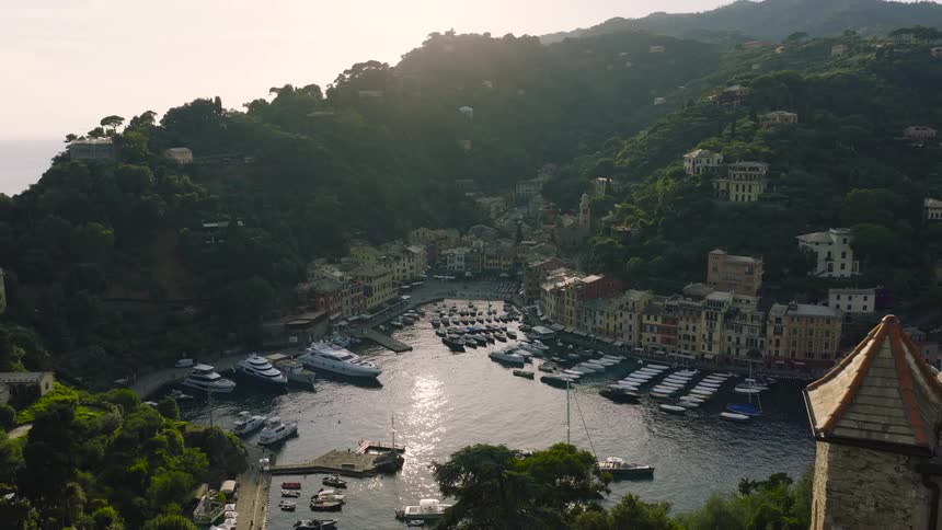 A stunning aerial shot reveals the picturesque Portofino harbor and its colorful village, flying past the historic Castello Brown at golden hour