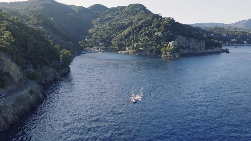 High angle aerial view of a boat sailing into a secluded bay surrounded by verdant green hills along the Italian Riviera