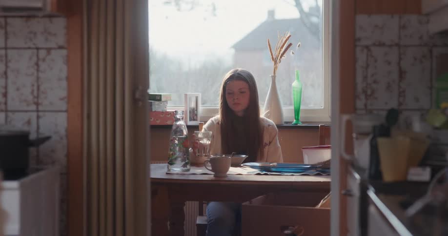A tired teenage girl slowly eats her breakfast at a sunlit kitchen table while her mother moves around in the foreground
