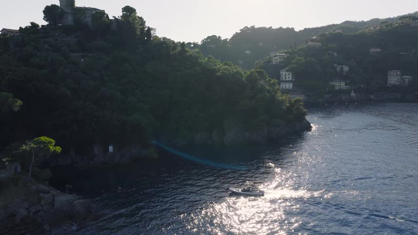 An aerial view of a small motorboat cruising along a beautiful, rocky coastline dotted with villas and a castle as the sun sparkles on the water