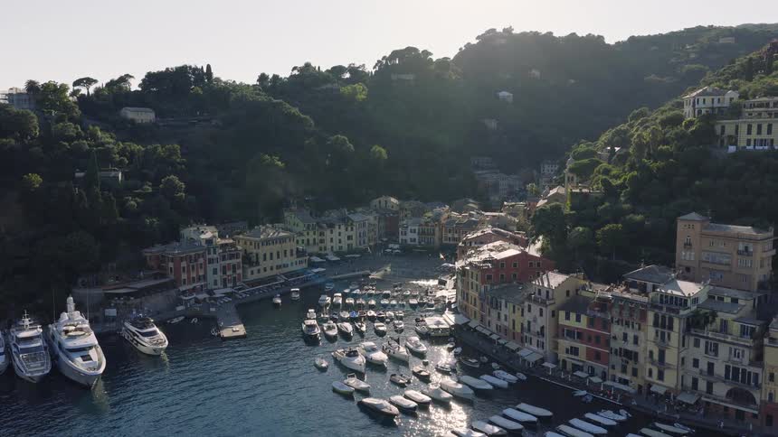 A stunning aerial reveal of the idyllic Portofino harbor, with its colorful buildings and sparkling bay filled with yachts, nestled in the green Italian Riviera