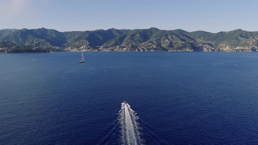An aerial drone follows a speed boat as it cruises across the deep blue sea toward a stunning mountainous coastline in the Mediterranean