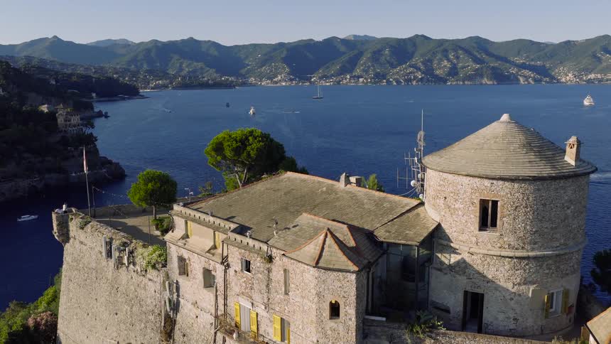 A scenic aerial view flies over the historic Castello Brown in Portofino, revealing the stunning blue waters of the Italian Riviera with boats dotting the sea