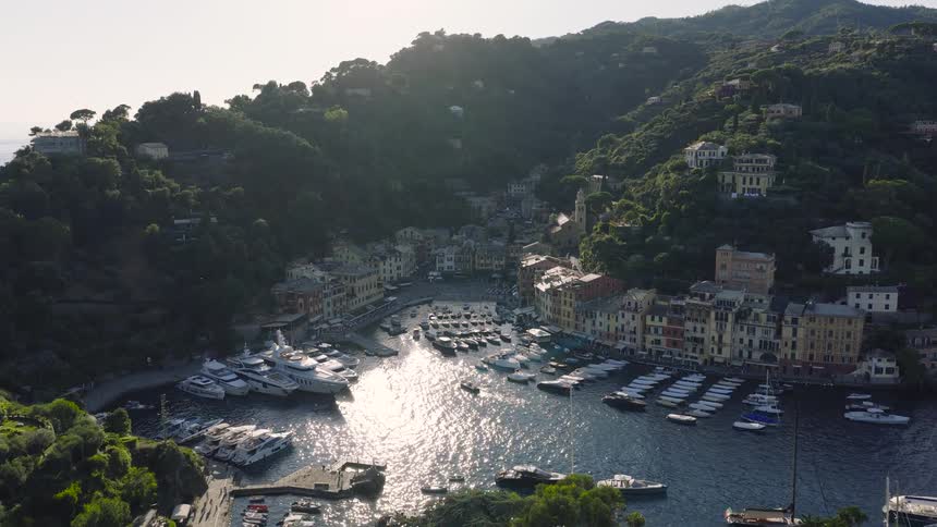 An aerial shot flies past a historic castle to reveal the beautiful sunlit harbor of Portofino, Italy, filled with boats and yachts
