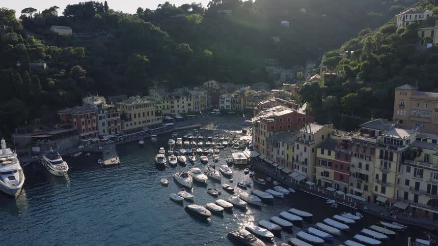 Stunning aerial shot flying towards the sun-drenched harbor of Portofino, Italy, revealing its colorful village and luxury yachts