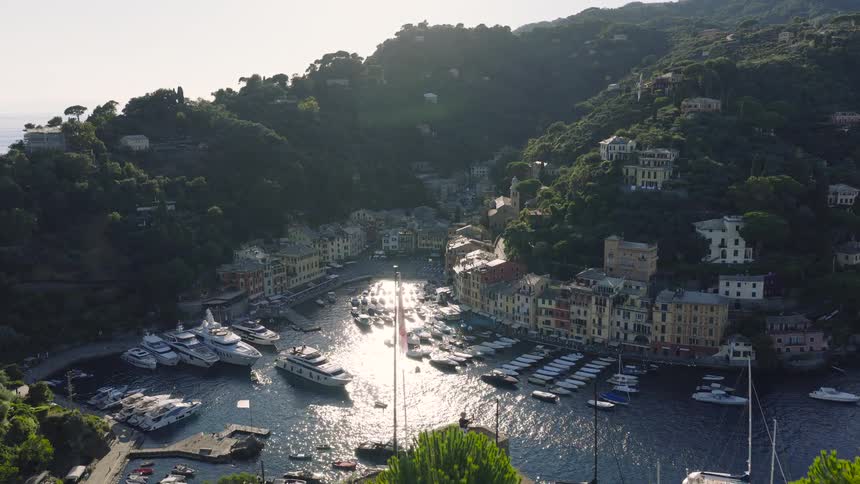 An aerial view flies over Castello Brown to reveal the stunning and iconic harbor of Portofino, Italy, filled with luxury yachts and boats