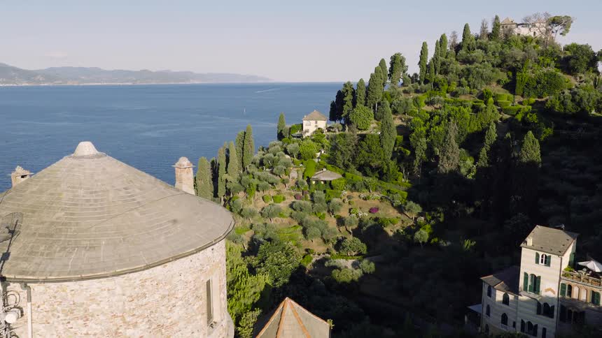 A scenic aerial flight over the historic Castello Brown and the lush, tree-covered cliffs of Portofino on the Italian Riviera