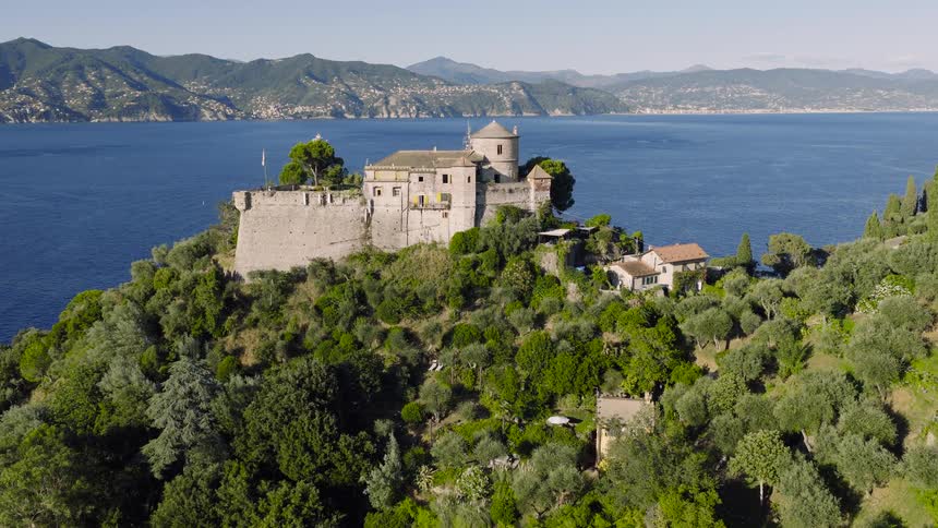 A stunning aerial view flies towards the majestic Castello Brown perched on a verdant cliff overlooking the beautiful blue sea of Portofino, Italy