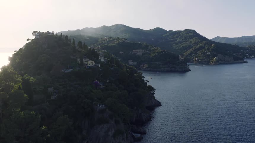 Stunning aerial footage flies over the iconic Portofino peninsula in Italy, showcasing the lighthouse and lush green hills dotted with villas at golden hour