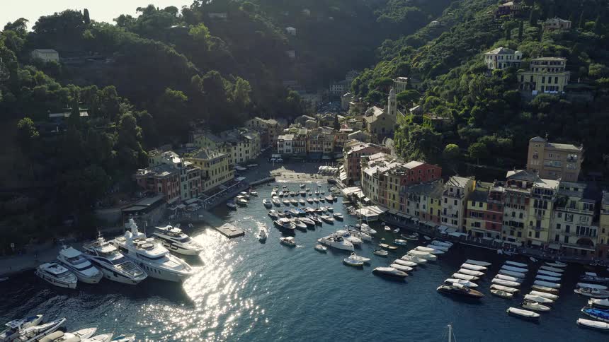 An aerial view flies over the historic Castello Brown to reveal the picturesque harbor of Portofino, Italy, filled with luxury yachts and boats