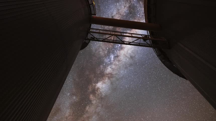 A dramatic upward-tilting timelapse reveals the magnificent Milky Way galaxy between two large observatory buildings at night