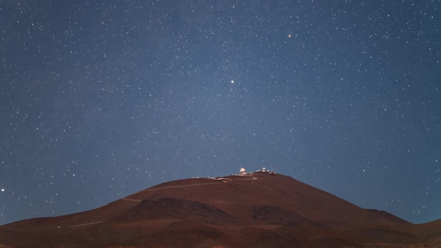 A stunning night timelapse shows stars trailing across the dark blue sky over a mountain-top astronomical observatory
