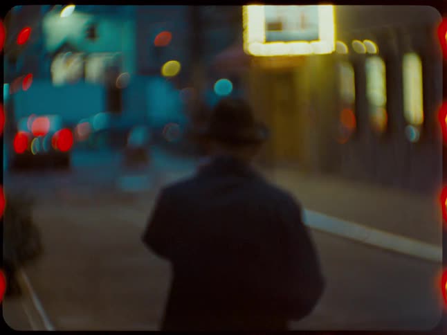 Dreamy, out-of-focus vintage film shot of a mysterious man in a hat walking down a city street at night