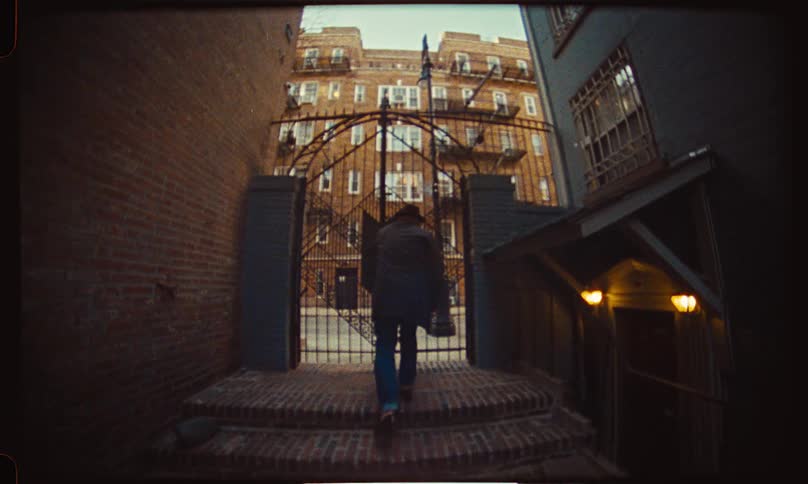 A mysterious man in a hat walks up steps in a city alleyway, opens a large iron gate, and exits onto the street in a vintage super 8mm film style
