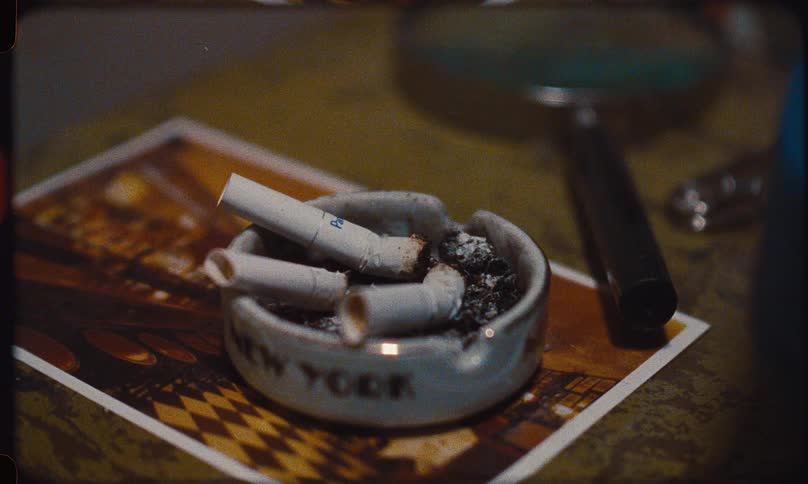 A gritty, vintage film shot captures a close-up of a New York souvenir ashtray overflowing with cigarette butts