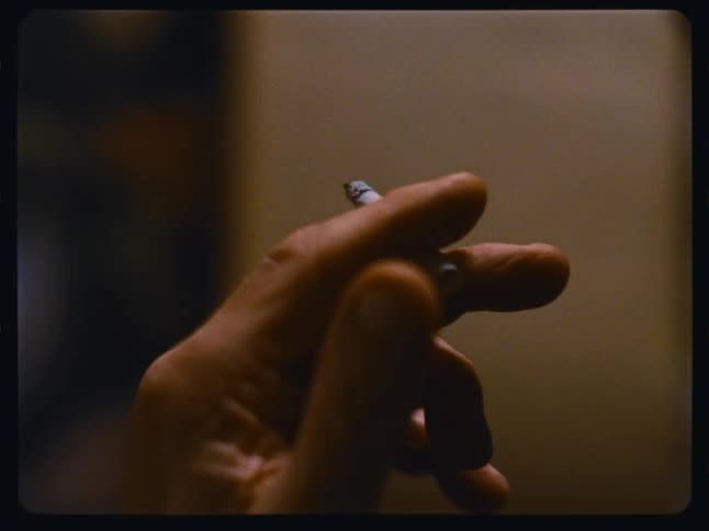 A close-up shot of a hand holding a lit cigarette in a dimly lit room, captured on vintage film