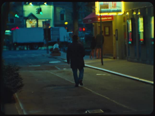 A man in a hat walks away from the camera down a dimly lit, gritty city street at night in a vintage film aesthetic
