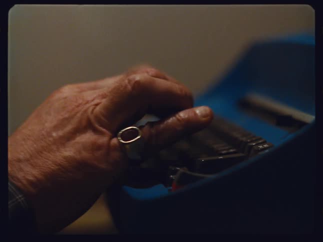 Close-up of an elderly person's hands with a large ring typing on a vintage blue typewriter in a warm, dimly lit room