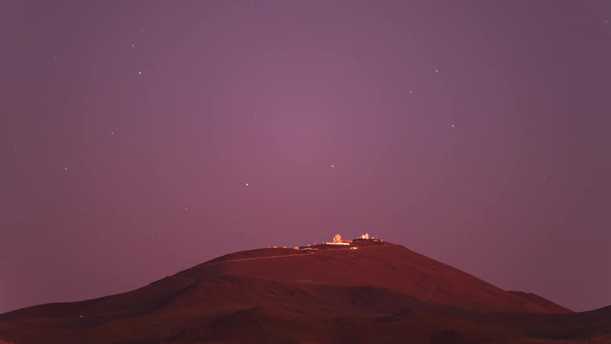 A day-to-night timelapse of the La Silla Observatory on a desert mountain, transitioning from a golden sunset to a vast, starry night sky with rotating constellations
