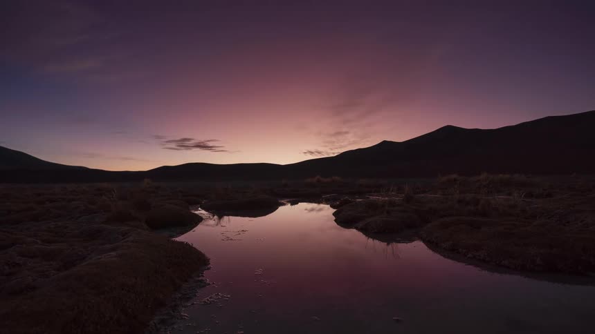 A stunning day-to-night timelapse of a sunset over a desert mountain landscape, with the colorful sky and stars reflecting in a still pool of water