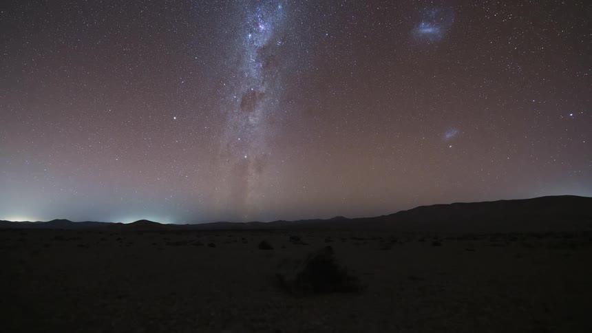A stunning day-to-night timelapse shows the Milky Way and moon rising over a vast, rocky desert after a colorful sunset