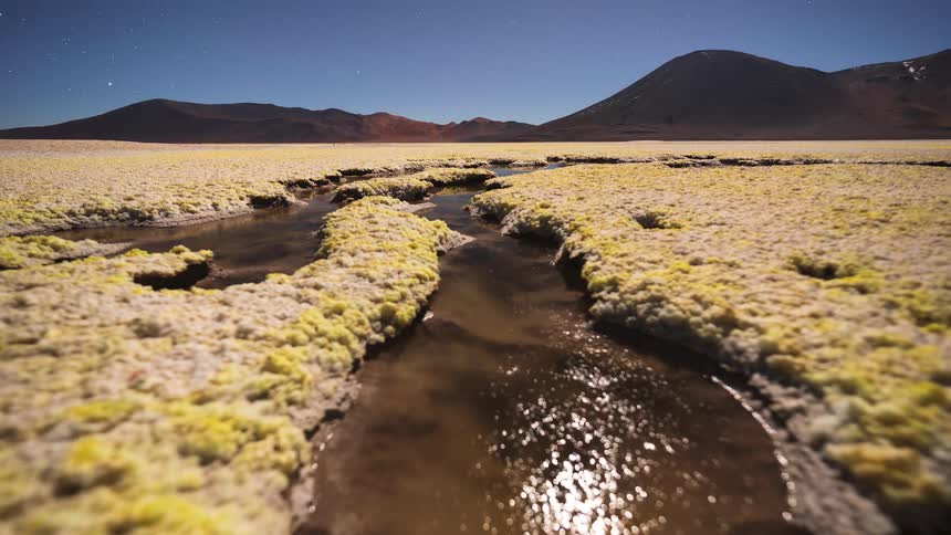 A low-angle night timelapse captures the rising moon and stars reflecting in a surreal, mineral-rich stream winding through a remote mountain landscape
