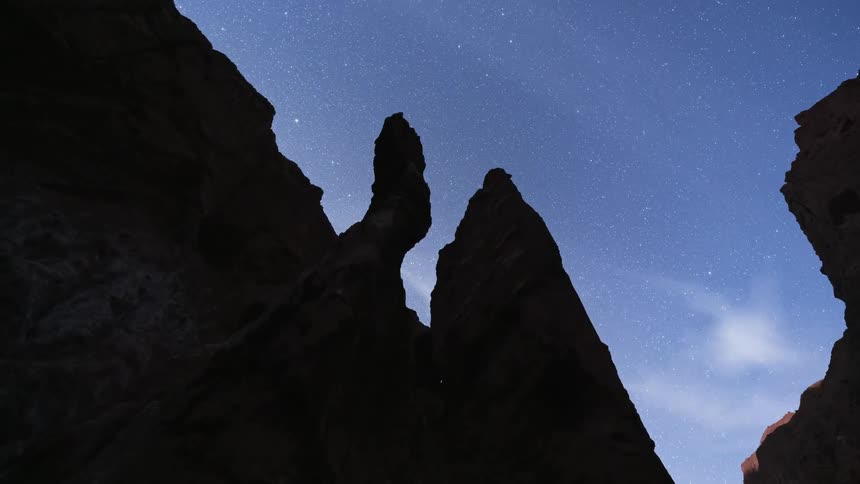 Awe-inspiring time-lapse captures the stars and Milky Way galaxy moving across the night sky above silhouetted rock formations, transitioning to dawn as the moon rises