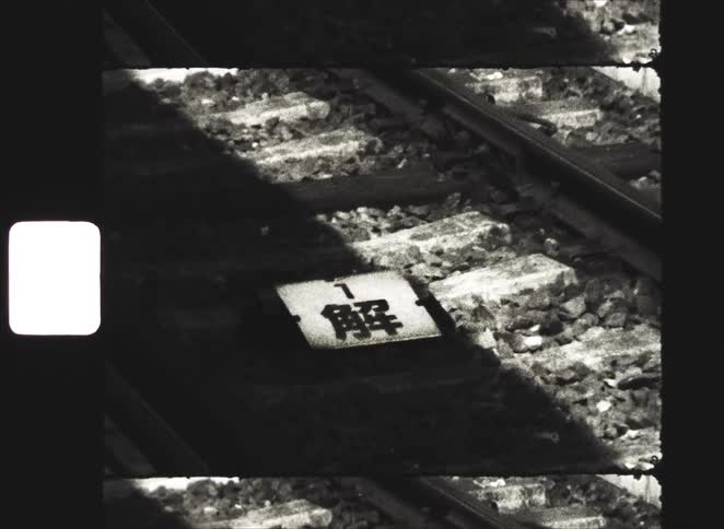 Gritty black and white Super 8 film footage shows a Japanese sign on a rocky railroad track