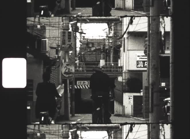Gritty, black and white 8mm film shows two people on bicycles riding away down a narrow city street in Japan