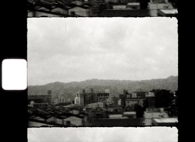 Vintage black and white 8mm film captures a tracking shot of a Japanese city with mountains in the distance