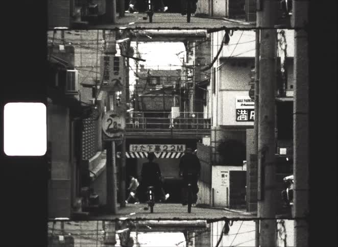 Vintage 8mm black and white footage shows two cyclists riding down a narrow, crowded alley in a Japanese city and into a dark underpass