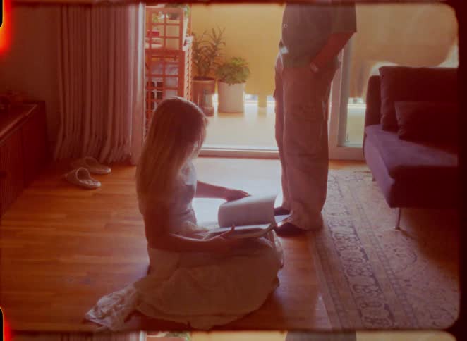 A nostalgic, sun-drenched film clip shows a woman reading a book on the floor while a man stands nearby in their apartment