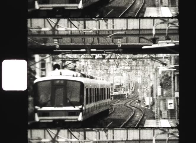 A vintage black and white film captures a train approaching on winding tracks through a dense urban landscape