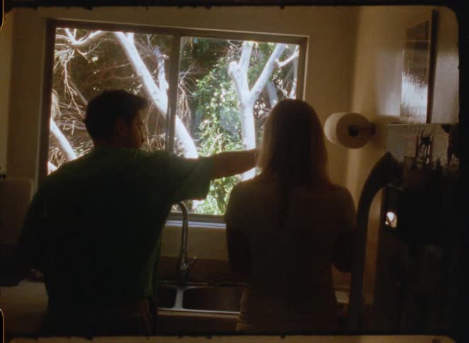 A silhouetted couple shares a tender moment while washing dishes in front of a sunny kitchen window, captured on vintage film