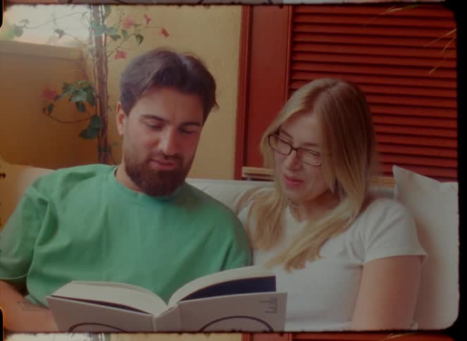 A happy couple sits close together on a couch, smiling as they read a book together in a warm, vintage-style shot
