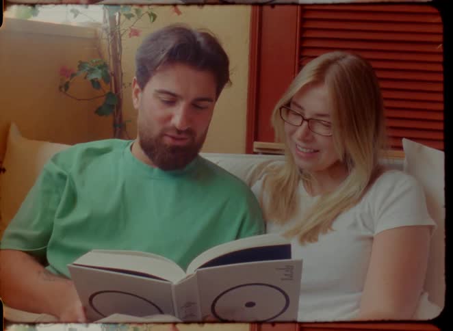 A relaxed young couple enjoys reading a book together in a sunlit room in this vintage-style footage