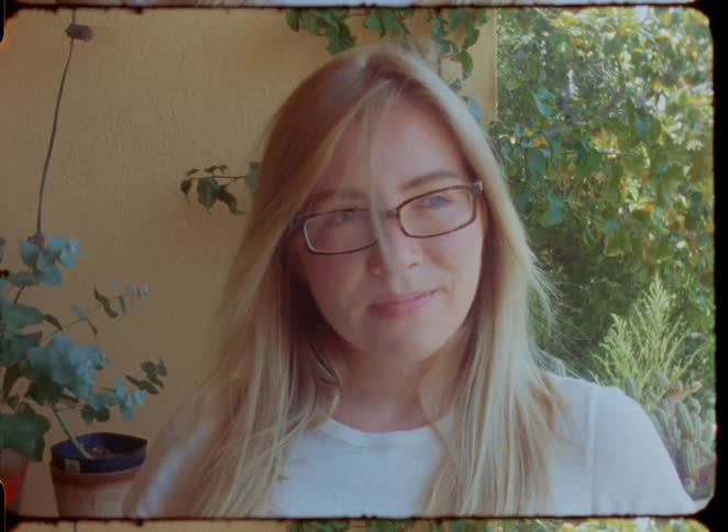 A vintage-style cinemagraph portrait of a young blonde woman with glasses smiling gently on a sunny patio
