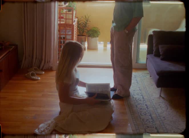A woman kneels on the floor of a sunlit living room, flipping through a book while a man stands nearby, captured in a vintage, film-like style