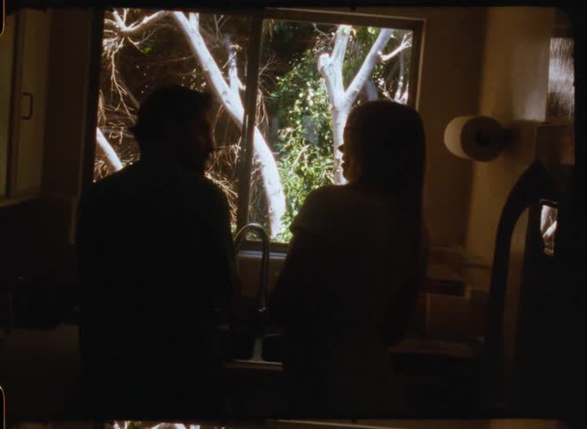 A silhouetted couple shares an intimate moment while washing dishes together in a warm, sunlit kitchen