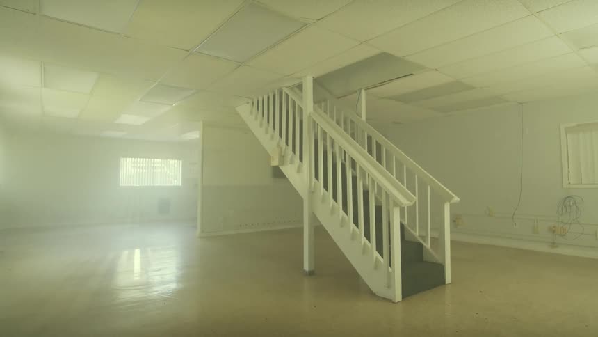 A slow tilt down from the ceiling reveals an empty, hazy basement with a central staircase illuminated by sunlight from the windows