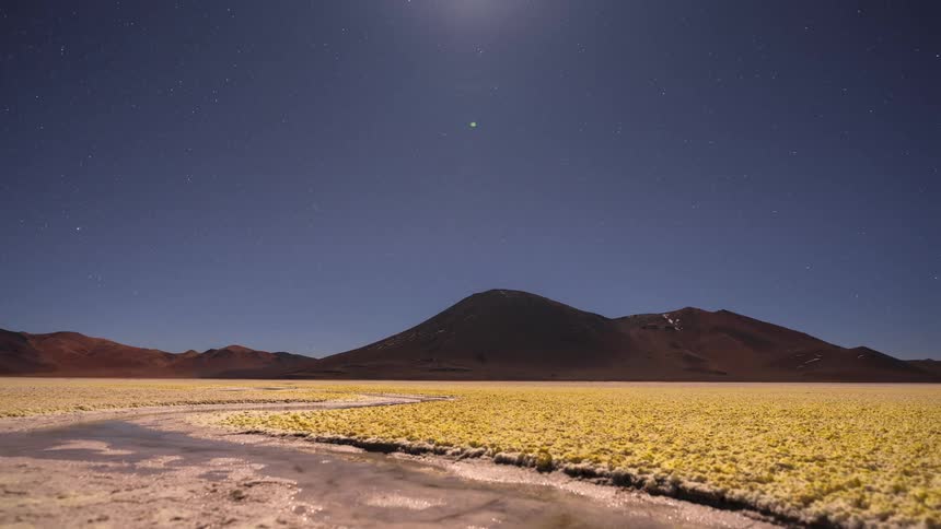 Celestial time-lapse of stars rotating and the moon rising over a surreal yellow salt flat and distant mountains