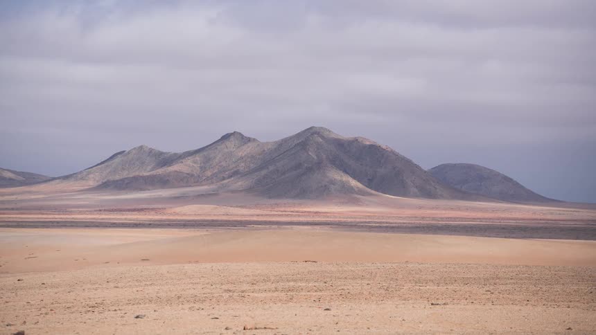 A serene time-lapse captures the shifting light and shadows over a vast, arid mountain range and desert floor