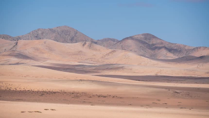 A time-lapse captures the subtle movement of shadows across a vast and arid desert landscape with sand dunes and distant mountains